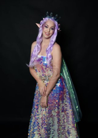 artistic portrait of beautiful female model with long purple hair and elf ears, wearing a fantasy fairy crown, wearing a rainbow glitter sequin ball gown. Standing in side profile, isolated on dark studio background.