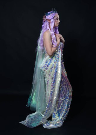 Retrato de cuerpo entero de la hermosa modelo femenina con el pelo largo de color púrpura con orejas de elfo, una corona de hadas de fantasía y vestido de lentejuelas arco iris brillante. pose elegante de pie, aislado en fondo oscuro estudio