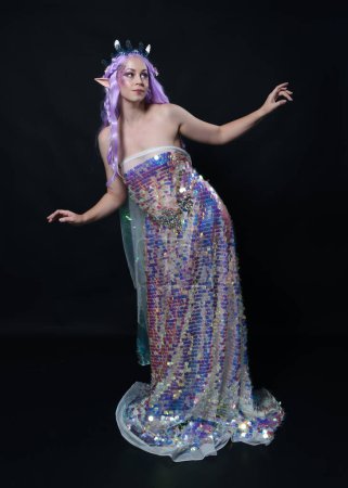 Photo for Full length portrait of beautiful female model with long purple hair wearing elf ears, a fantasy fairy crown and  rainbow glitter sequin ball gown. graceful standing pose,  isolated in dark studio background - Royalty Free Image