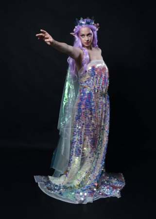 Retrato de cuerpo entero de la hermosa modelo femenina con el pelo largo de color púrpura con orejas de elfo, una corona de hadas de fantasía y vestido de lentejuelas arco iris brillante. pose elegante de pie, aislado en fondo oscuro estudio
