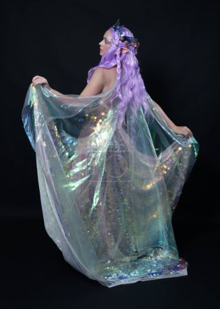 Photo for Full length portrait of beautiful female model with long purple hair wearing elf ears, a fantasy fairy crown and  rainbow glitter sequin ball gown.  walking away, back view, isolated dark studio - Royalty Free Image