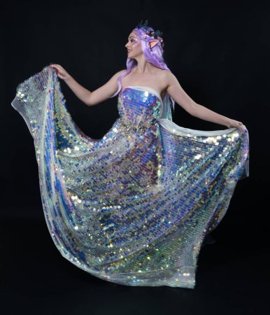 Full length portrait of beautiful female model with long purple hair wearing elf ears, a fantasy fairy crown and  rainbow glitter sequin ball gown. graceful standing pose,  isolated in dark studio background