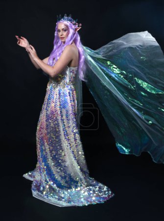 Photo for Full length portrait of beautiful female model with long purple hair wearing elf ears, a fantasy fairy crown and  rainbow glitter sequin ball gown.  walking away, back view, isolated dark studio - Royalty Free Image
