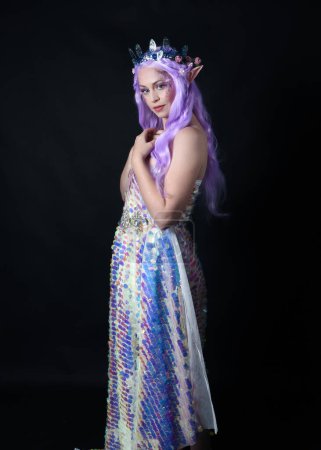 Photo for Full length portrait of beautiful female model with long purple hair wearing elf ears, a fantasy fairy crown and  rainbow glitter sequin ball gown. graceful standing pose,  isolated in dark studio background - Royalty Free Image