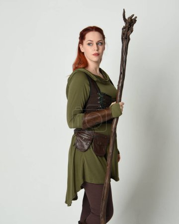  Porträt eines schönen rothaarigen weiblichen Modells, das ein grünes mittelalterliches Fantasiekostüm mit brauner Rüstung trägt. Stehende Pose mit Holzzauberstab Waffe, isolierter weißer Studiohintergrund.