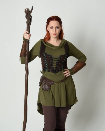  Porträt eines schönen rothaarigen weiblichen Modells, das ein grünes mittelalterliches Fantasiekostüm mit brauner Rüstung trägt. Stehende Pose mit Holzzauberstab Waffe, isolierter weißer Studiohintergrund.