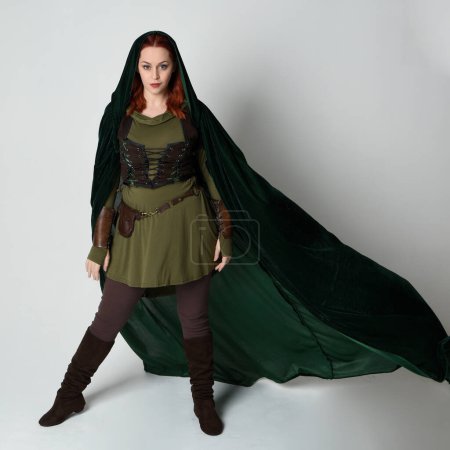 Ganztägiges Porträt eines rothaarigen weiblichen Modells in einem grünen mittelalterlichen Kostüm aus Leder und einem dramatischen, lang fließenden Kapuzenumhang aus Samt. Standing Walking Pose, isolierter weißer Studiohintergrund.