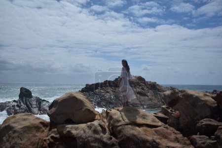 retrato de modelo femenino de pie vistiendo vestido de diosa blanca, dramático paisaje natural de la costa del océano rocoso con acantilados de piedra. castle rock, Busselton, Australia Occidental