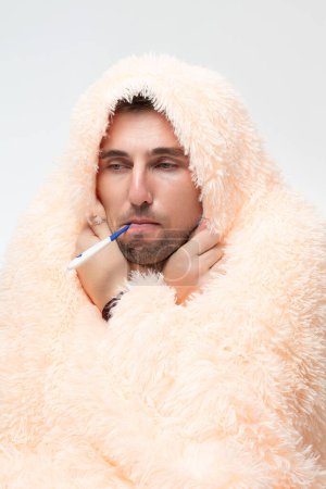 Foto de A sick man wrapped in a blanket holds a thermometer in his mouth, healthcare and treatment. - Imagen libre de derechos