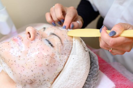 Téléchargez les photos : A beautician removes a moisturizing sheet mask on a woman's face with a spatula, cosmetology procedures. - en image libre de droit