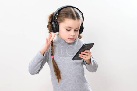Photo for A girl with headphones and a phone on a white background, a girl with pigtails listens to music in headphones, photo with copy space, wireless headphones and a child with a smartphone. - Royalty Free Image