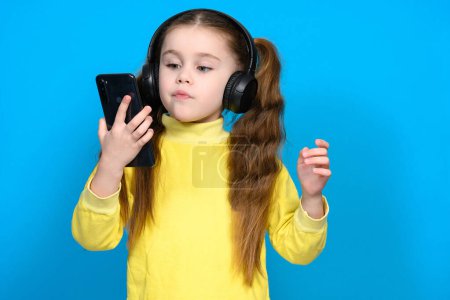 Photo for Portrait of a little girl with a phone and wireless headphones, a child in a yellow sweater on a blue background, a child dances when he hears music. - Royalty Free Image