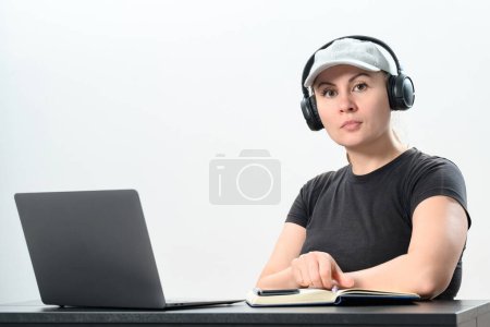 Photo for Busy young woman in headphones working on laptop, making video call and making notes in notebook, work and study online, copy space and white background. - Royalty Free Image