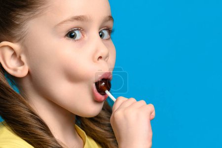 Auf blauem Hintergrund leckt ein Mädchen einen runden Lutscher in Großaufnahme, Karies und schlechtes Futter für die Zähne.