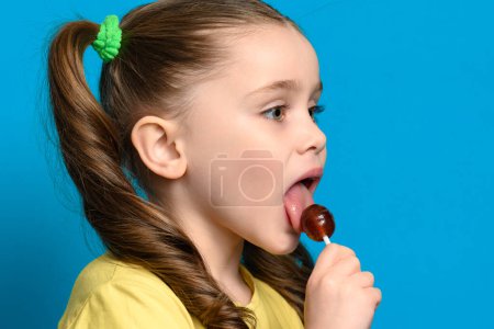 Photo for On a blue background, a girl licks a round lollipop close-up, caries and bad food for teeth. - Royalty Free Image