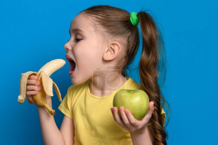 Green apple and banana are held by a child on a blue background, a girl for healthy and healthy food, blue background and copy space.