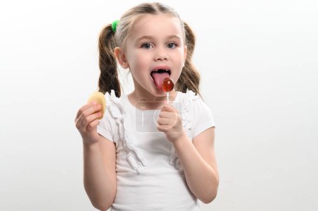 A little girl licks a lollipop and holds potato chips in her hand on a white background, baby teeth and junk food.