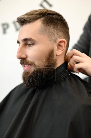 Foto de Un joven con una barba en una capa negra se sienta en una silla en una barbería. El cliente está esperando un corte de pelo y el afeitado de la barba. - Imagen libre de derechos