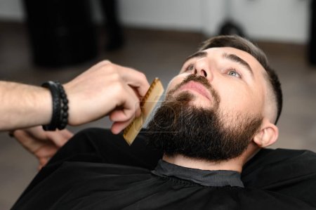 Photo for Barber combs beard with comb while cutting and shaving Caucasian man - Royalty Free Image