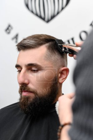 Foto de Un barbero corta a un hombre con barba en una peluquería. Corte de pelo corto del cliente con un clipper. - Imagen libre de derechos