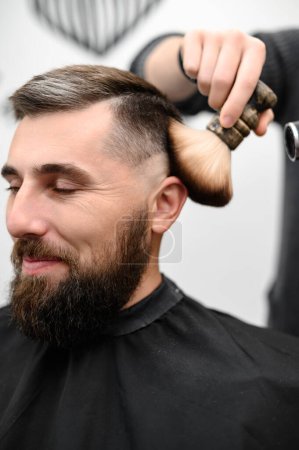 Foto de Peluquero barre el cabello con un cepillo mientras corta el cabello de un cliente en un salón. - Imagen libre de derechos