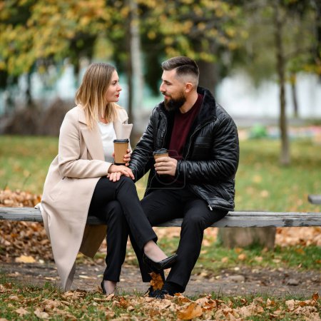Foto de Una pareja, un hombre y una mujer, están sentados en el parque en un banco con café en sus manos, una reunión en el parque. - Imagen libre de derechos