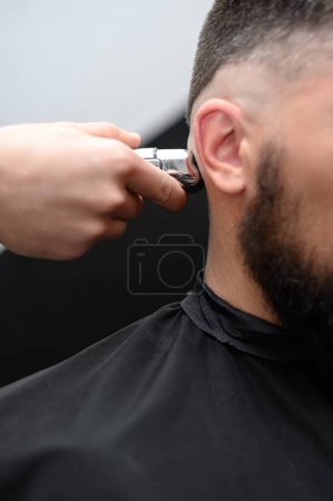 Peluquero afeita la sien con un recortador inalámbrico durante un corte de pelo corto en los lados de la cabeza.