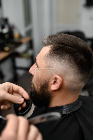 Foto de Peluquero barre el cabello con un cepillo mientras corta el cabello de un cliente en un salón. - Imagen libre de derechos
