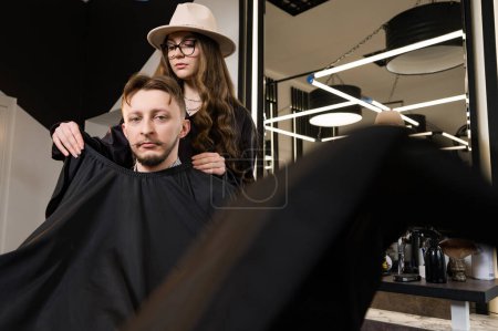 Foto de Un joven con una barba en una capa negra se sienta en una silla en una barbería. El cliente está esperando un corte de pelo y el afeitado de la barba. - Imagen libre de derechos