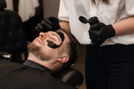 Während der Rasur trägt Barber eine schwarze feuchtigkeitsspendende Maske auf das Gesicht des Mannes auf. Ein Dermatologe trägt eine schwarze Maske auf das Gesicht eines bärtigen Patienten auf, um Poren und Falten zu reinigen.