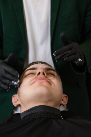 A barber in black gloves applies a moisturizing white cream to a clients face. A beautician applies a white mask with a vitamin complex to the skin of a mans face.