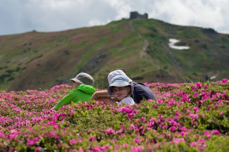 Three children sat down to rest on a lawn with rhododendrons against the background of Mount Pip Ivan, summer active recreation for children.