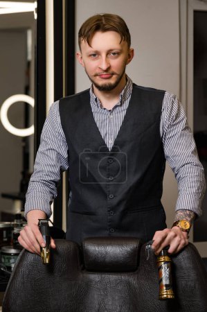 Foto de Un peluquero con camisa y chaleco está cerca de una silla de peluquero sosteniendo herramientas para cortes de pelo. - Imagen libre de derechos