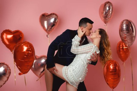 Photo for A couple in evening dresses are kissing while dancing against a background of red and pink balloons in the shape of a heart on a pink background. Vespers on Valentine's Day or March 8, Women's Day - Royalty Free Image