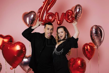 Photo for Couple in black suits holding love inscription, red balloons, pink background. A woman in a corset and jewelry with a man. They celebrate a party in honor of Valentine's Day - Royalty Free Image
