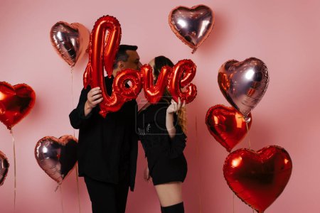Photo for Couple in black suits holding love inscription, red balloons, pink background. They celebrate a party in honor of Valentine's Day, post Card kiss - Royalty Free Image
