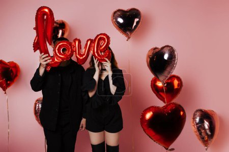Photo for Couple in black suits holding love inscription, red balloons, pink background. They celebrate a party in honor of Valentine's Day, post Card - Royalty Free Image