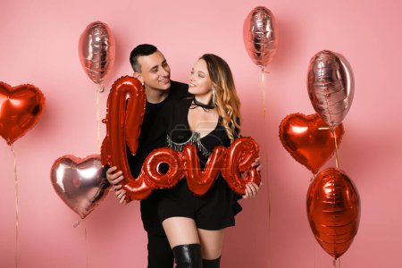 Photo for Couple in black suits holding love inscription, red balloons, pink background. A woman in a corset and jewelry with a man. They celebrate a party in honor of Valentine's Day - Royalty Free Image