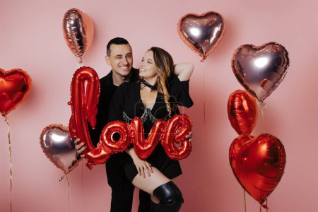 Photo for Couple in black suits holding love inscription, red balloons, pink background. Celebrate Valentine's Day party, dancing and fun - Royalty Free Image