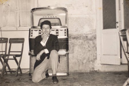 Foto de Un joven fumando y posando frente a una máquina de discos. Italia, 1960, archivo, fotografía en blanco y negro. - Imagen libre de derechos