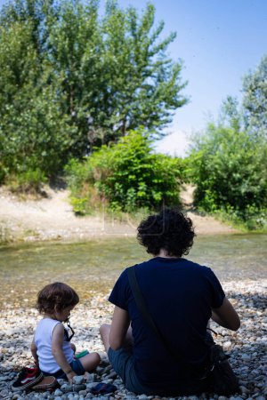 Foto de Joven padre e hijo pequeño bebé, vista trasera, sentado en la playa del río en un hermoso día soleado. Parco del Ticino, Lombardía, Italia. - Imagen libre de derechos