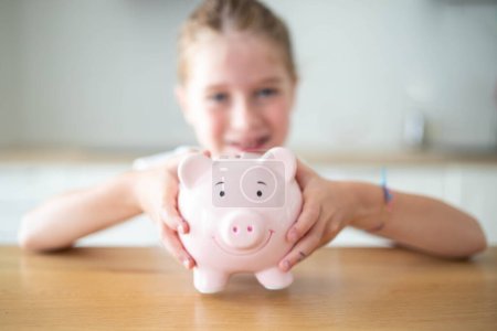 Photo for Caucasian child girl holding a smiling pink piggy bank in her hands on a wooden table. Saving money for kids concept. - Royalty Free Image