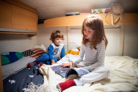 Foto de Dos niños caucásicos, hermano y hermana, leyendo libros en pijama en una cama de autocaravana durante una parada de viaje por carretera. Camper vacaciones viajando con niños. - Imagen libre de derechos