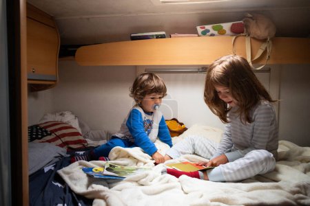 Téléchargez les photos : Deux enfants caucasiens, frère et sœur, lisant des livres en pyjama sur un lit de camping-car lors d'un arrêt sur la route. Vacances en camping-car avec enfants. - en image libre de droit