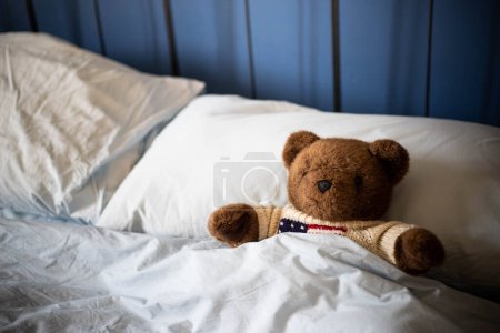 Brown teddy bear, plush toy, laying under covers in parents' bed with blue blankets and soft day light. Side view, background with copy space..