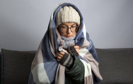 Téléchargez les photos : Une femme enveloppée dans une couverture se réchauffe alors qu'elle est assise dans une chambre froide en hiver. - en image libre de droit