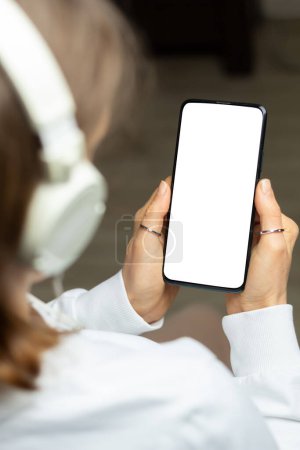 Téléchargez les photos : Une femme dans un casque tient un téléphone avec un écran blanc écoutant de la musique. Modèle de smartphone. - en image libre de droit