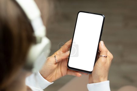 Téléchargez les photos : Une femme dans un casque tient un téléphone avec un écran blanc écoutant de la musique. Modèle de smartphone. - en image libre de droit