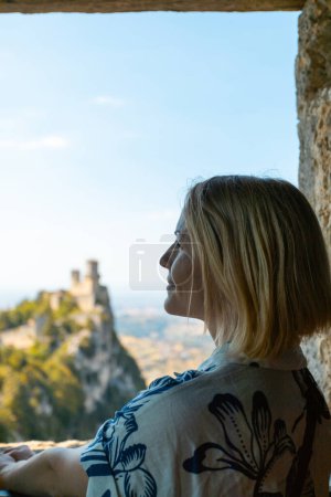 Téléchargez les photos : Young beautiful woman visits the sights of the Republic of San Marino, Italy. She stay near the window in Second tower - Cesta and admiring the view from the window on the panorama of San Marino and - en image libre de droit