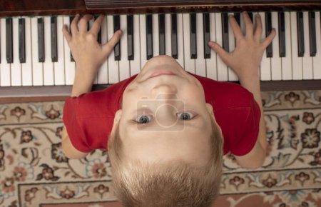 Téléchargez les photos : Jeune garçon assis au piano à la maison. Le garçon tient ses mains sur les clés et lève les yeux - en image libre de droit